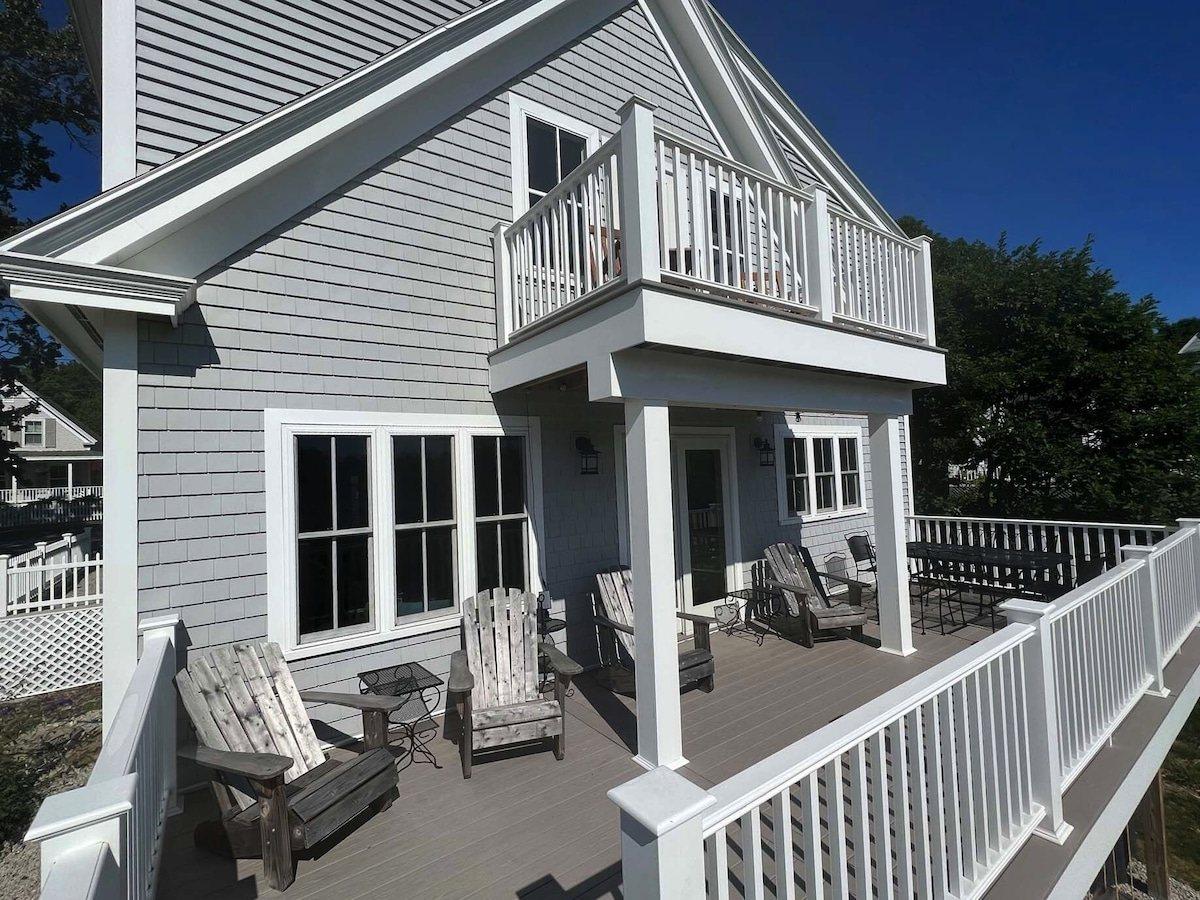 Main deck along the back of the house with Adirondack chairs, a dining table, and a small upper balcony above.