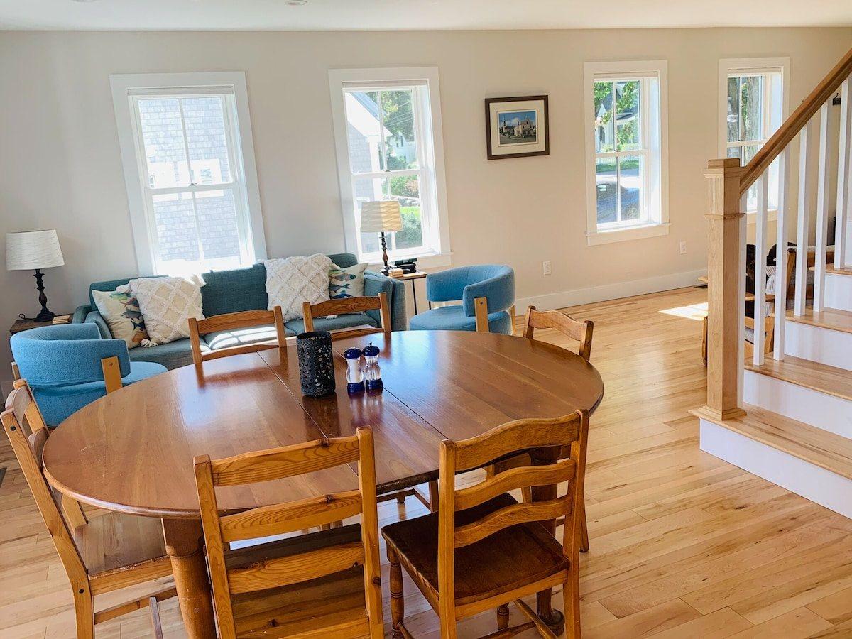 Round wooden dining table beside a sofa and two blue armchairs, with the staircase to the upper floor at right.