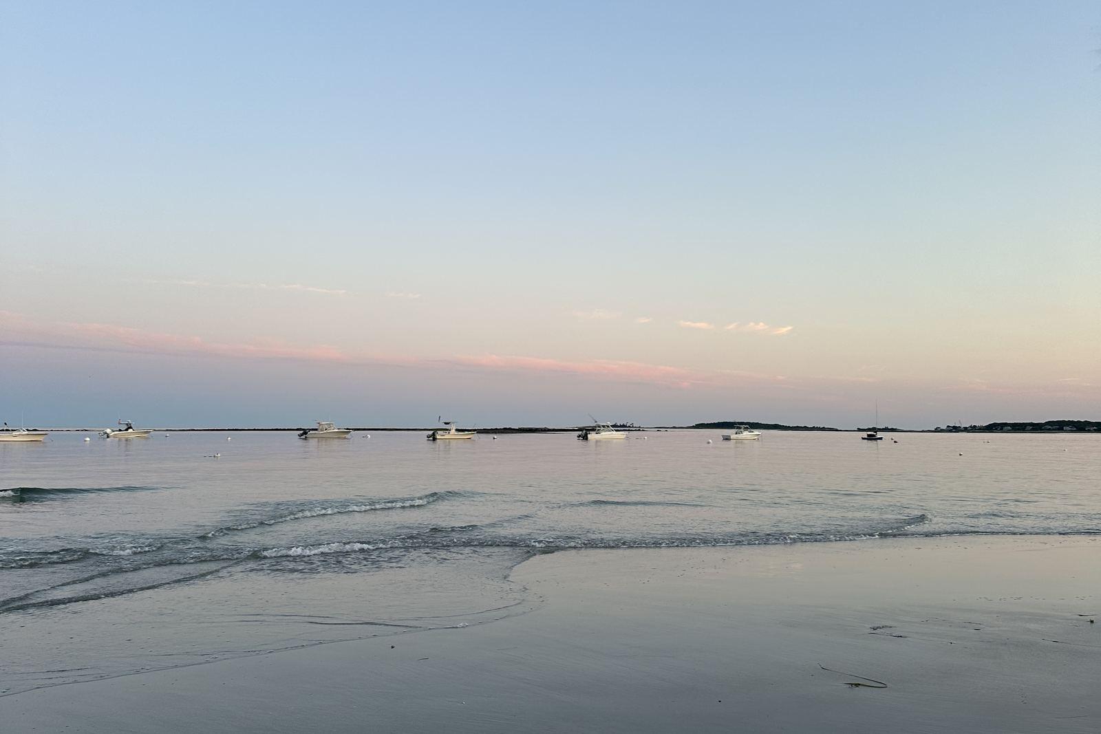 Small boats moored in calm water off Goose Rocks Beach at sunrise, with wet sand in the foreground and a pale pink sky.