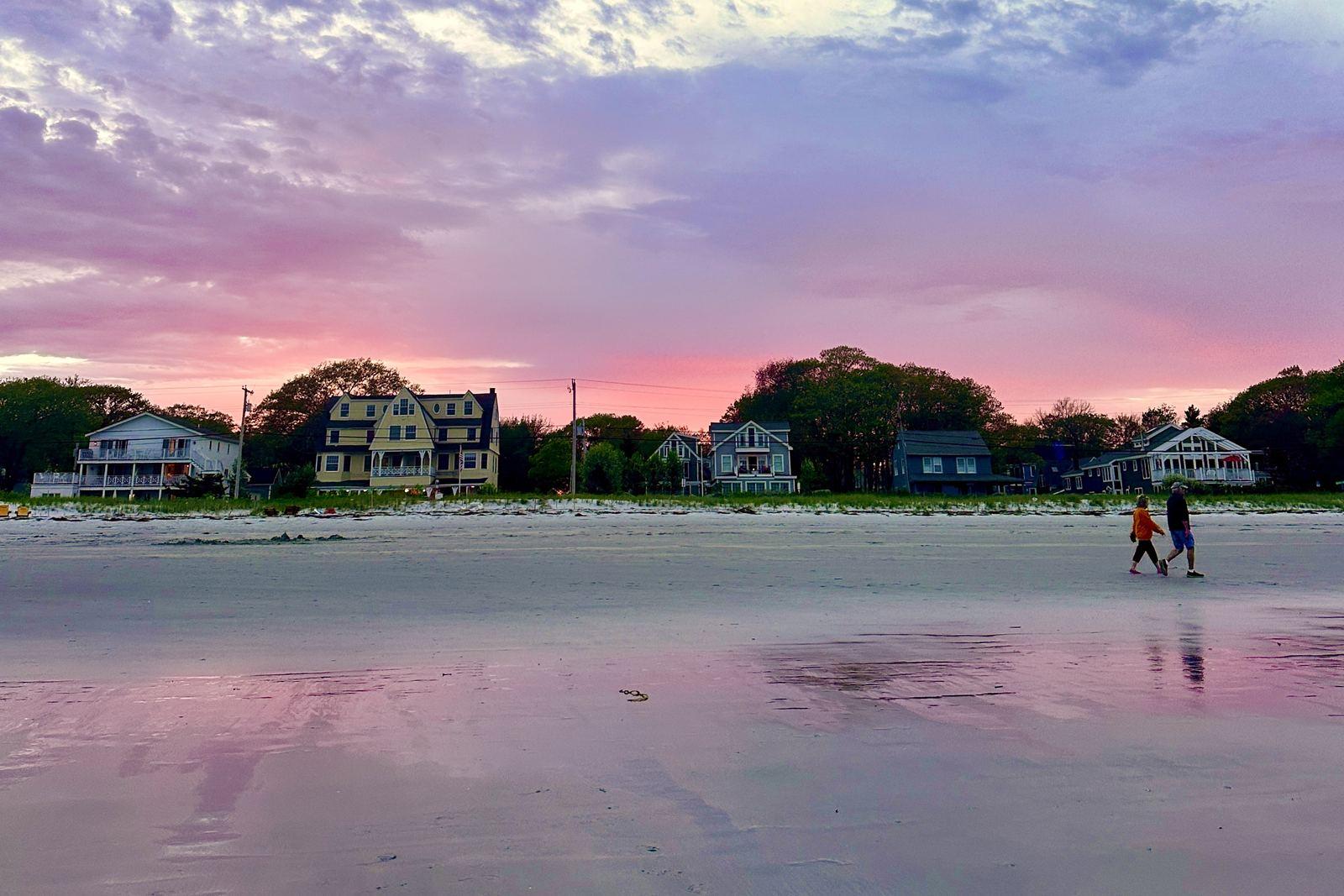 Two people walking along the wet sand at Goose Rocks Beach at sunset, with houses and a pink-purple sky behind them.
