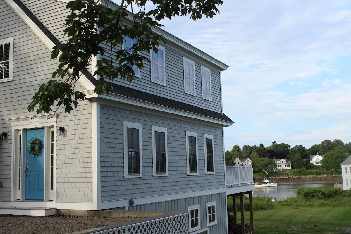 The gray-shingled house from the side: two floors of windows, white trim, a blue door with a wreath, and the deck at the far end, with Cape Porpoise harbor and a lobster boat behind it.