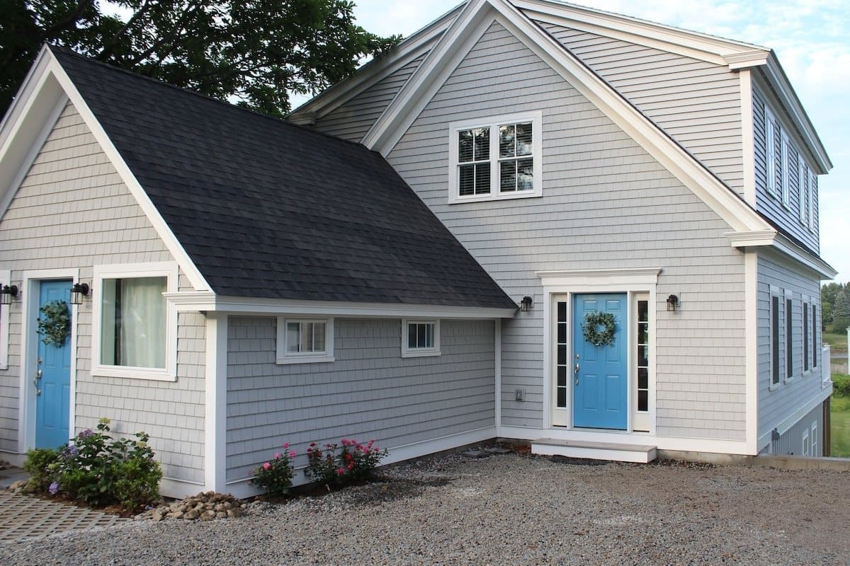 Front of the house from the gravel drive: two blue doors, white trim, gray shingles, roses along the foundation.
